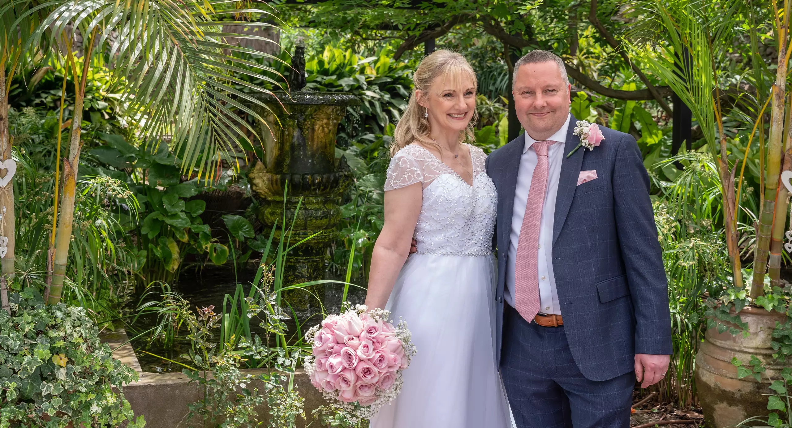 A bride in a white dress holding a bouquet of pink roses stands next to a groom in a suit with a pink tie, posing outdoors in a lush garden.
