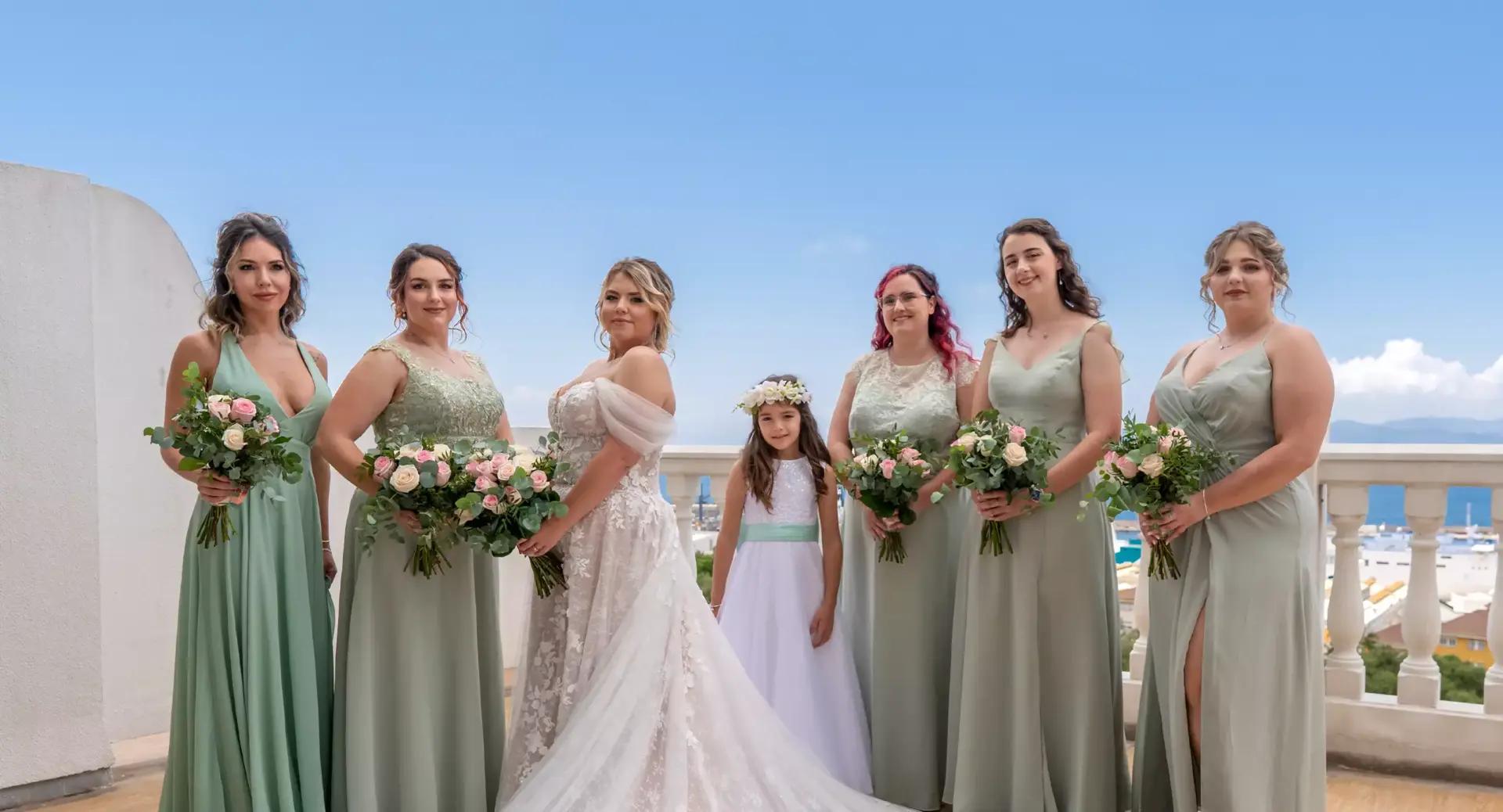 A bride in a white gown stands with six bridesmaids in sage green dresses and a flower girl in white, all holding bouquets, against a bright outdoor backdrop.