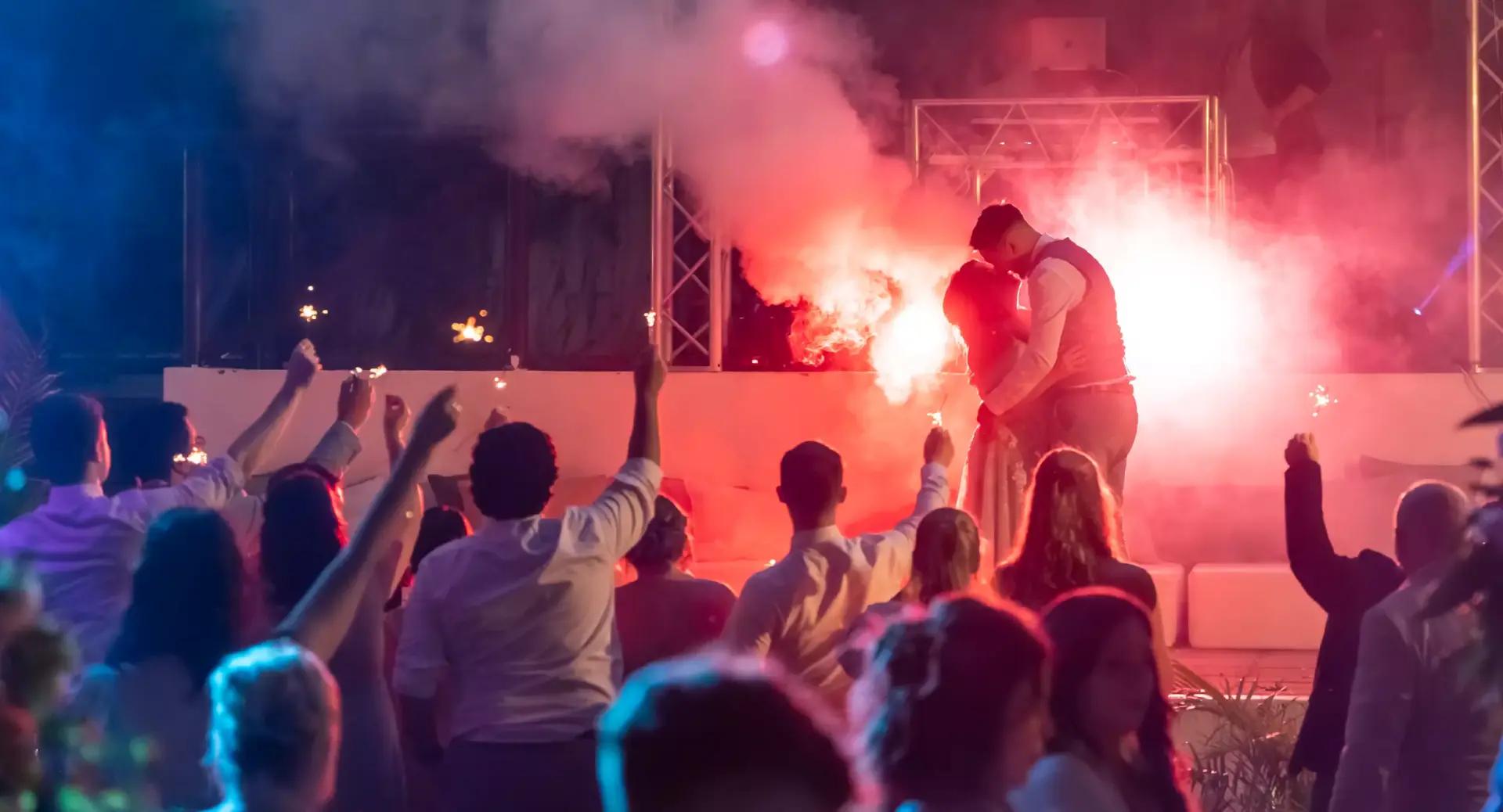 A couple embraces and kisses on stage under bright lights and smoke, while a crowd of people holding sparklers watches and raises their arms.