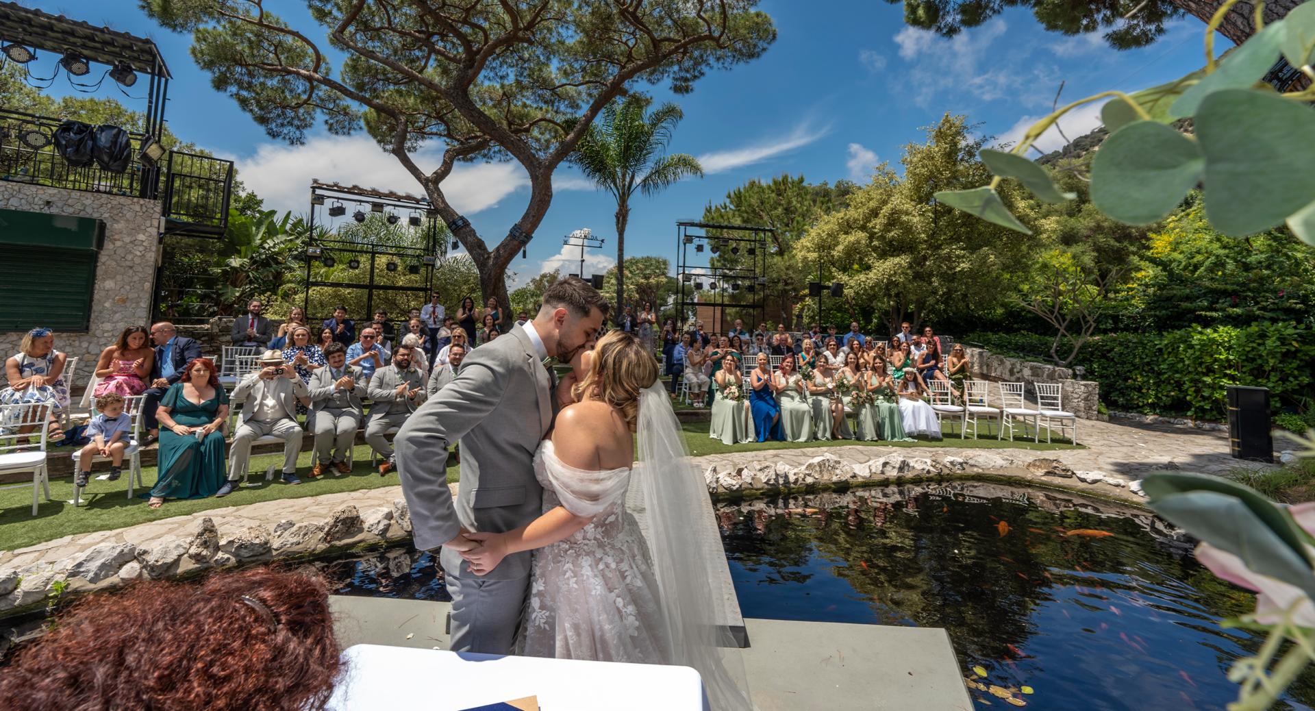 A bride and groom kiss at an outdoor wedding ceremony by a small pond, with guests seated and watching in the background under a sunny sky.