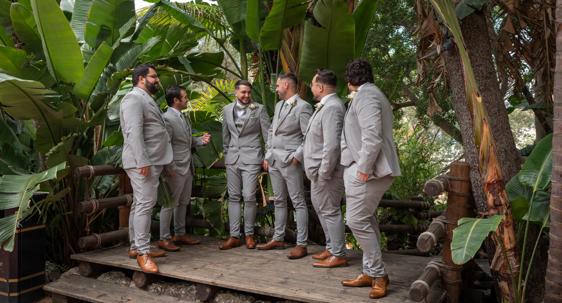 Six men in matching light gray suits and brown shoes stand together and talk on a wooden platform surrounded by lush green plants and trees.