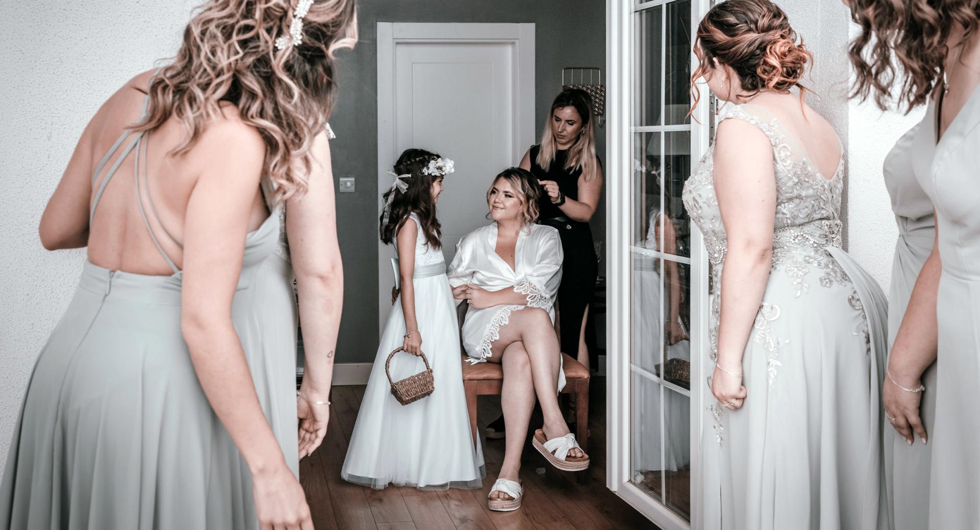 A bride in a white robe sits on a chair, smiling at a flower girl, while bridesmaids in pale dresses stand around them in a hallway.