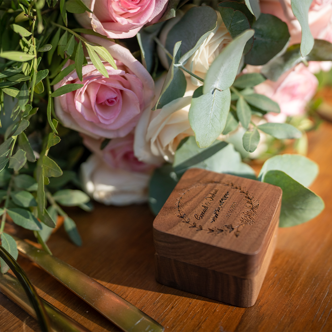 A wooden ring box with engraved text sits on a wooden table next to a bouquet of pink and white roses and green foliage.