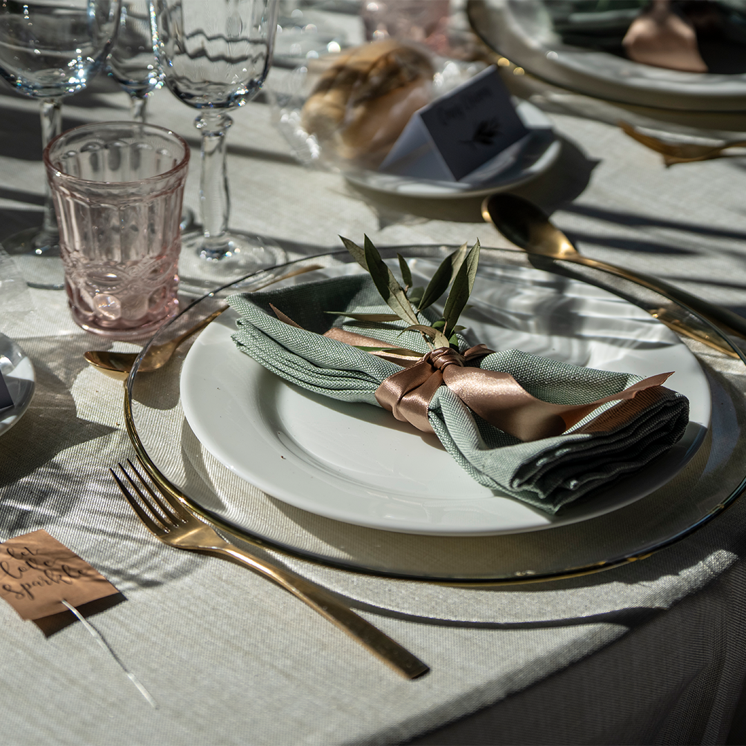 A formal table setting with a white plate, gold cutlery, green napkin tied with a ribbon, and a sprig of greenery on a beige tablecloth, with glassware and bread in the background.