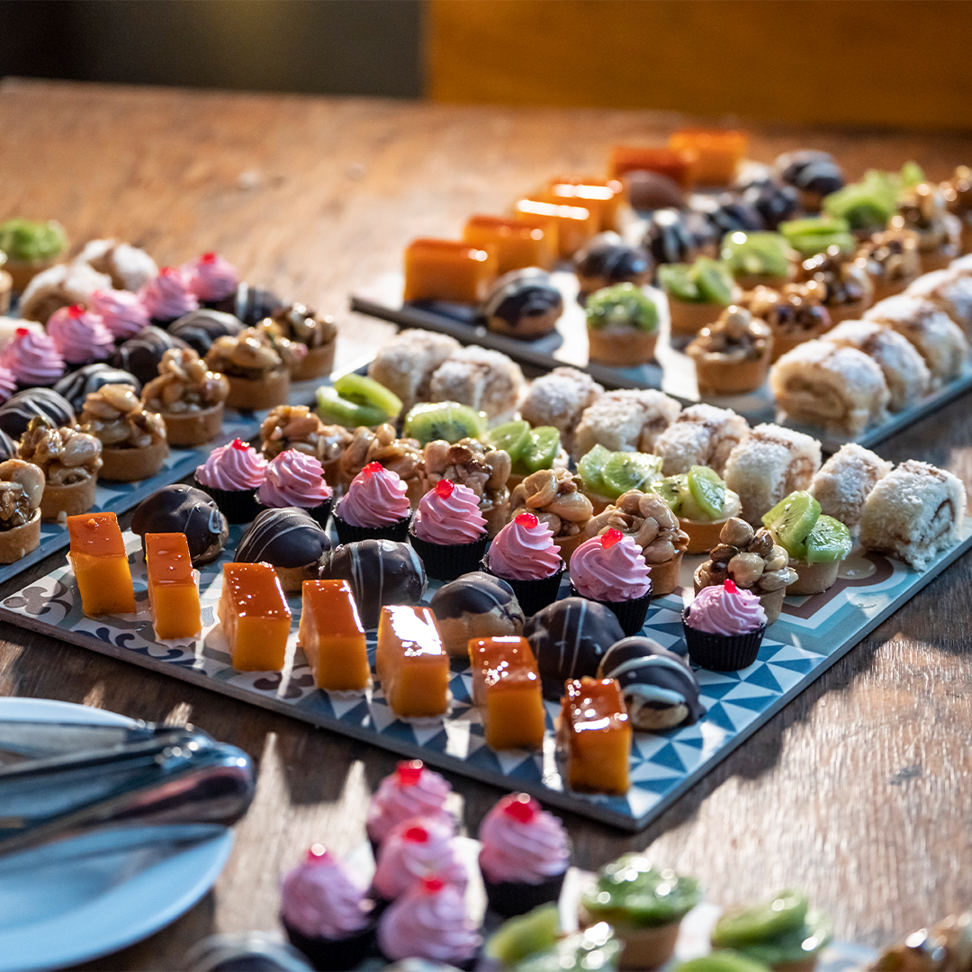 An assortment of colorful bite-sized desserts, including pastries, cakes, and sweets, arranged neatly on trays atop a wooden table.
