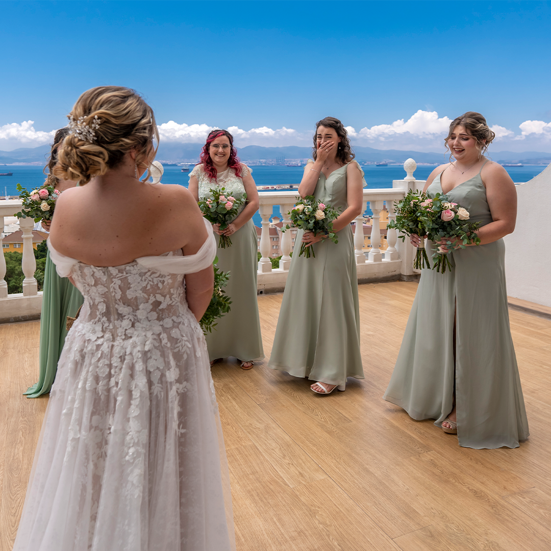 A bride in a white lace gown stands facing four bridesmaids in light green dresses holding bouquets on a balcony with a scenic ocean view.