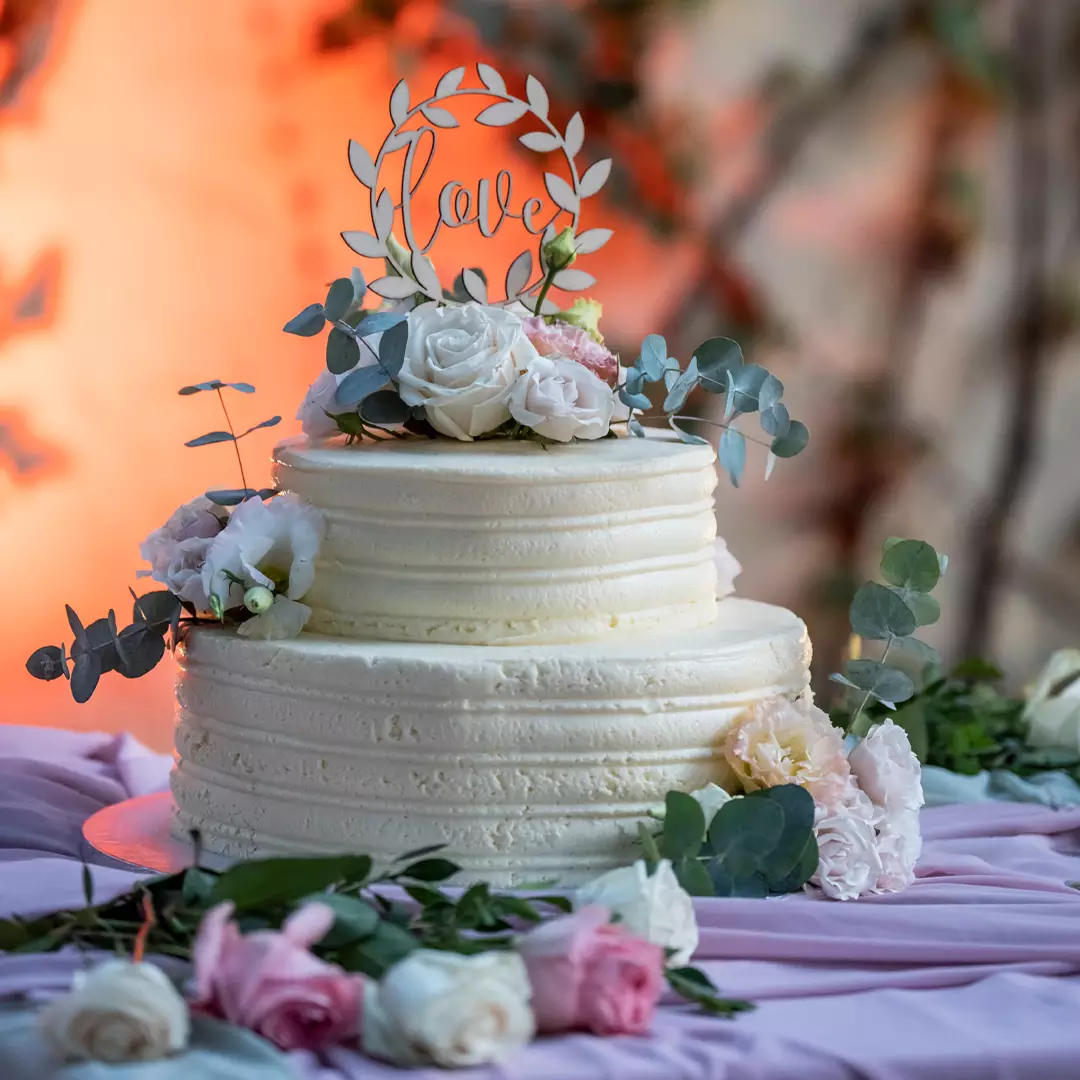 A two-tier white frosted cake decorated with roses, eucalyptus leaves, and a 