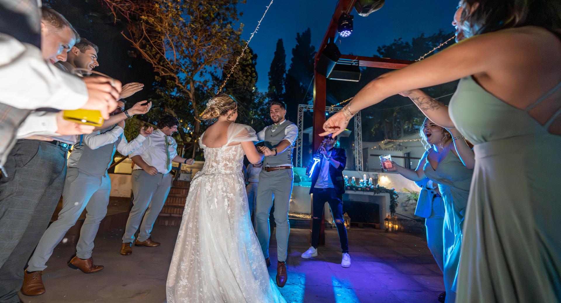 A bride and groom dance surrounded by guests at an outdoor evening wedding reception, illuminated by string lights and blue stage lighting.