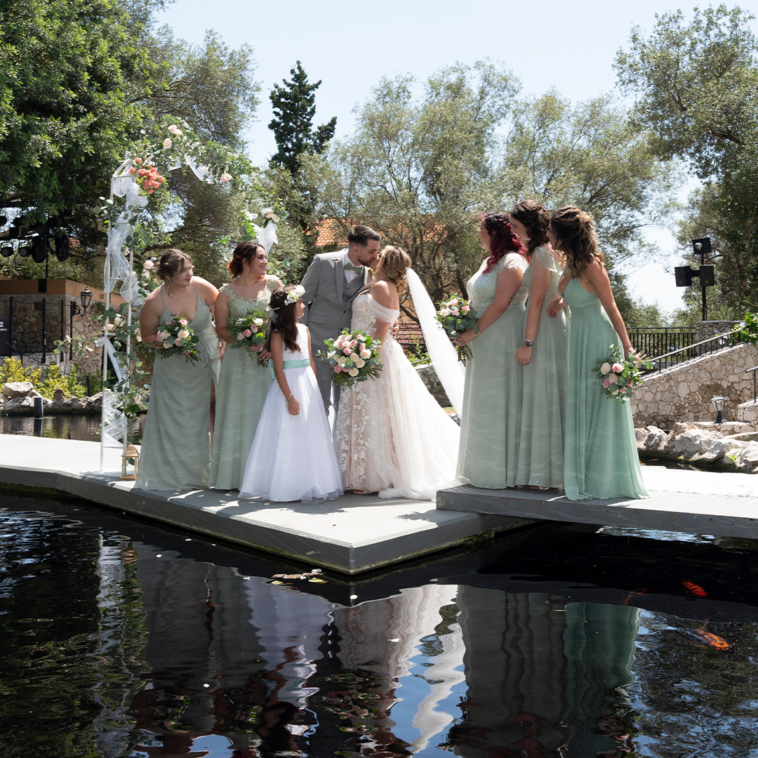 A bride and groom stand with six bridesmaids on a dock by the water, all holding bouquets and dressed in formal attire under daylight.