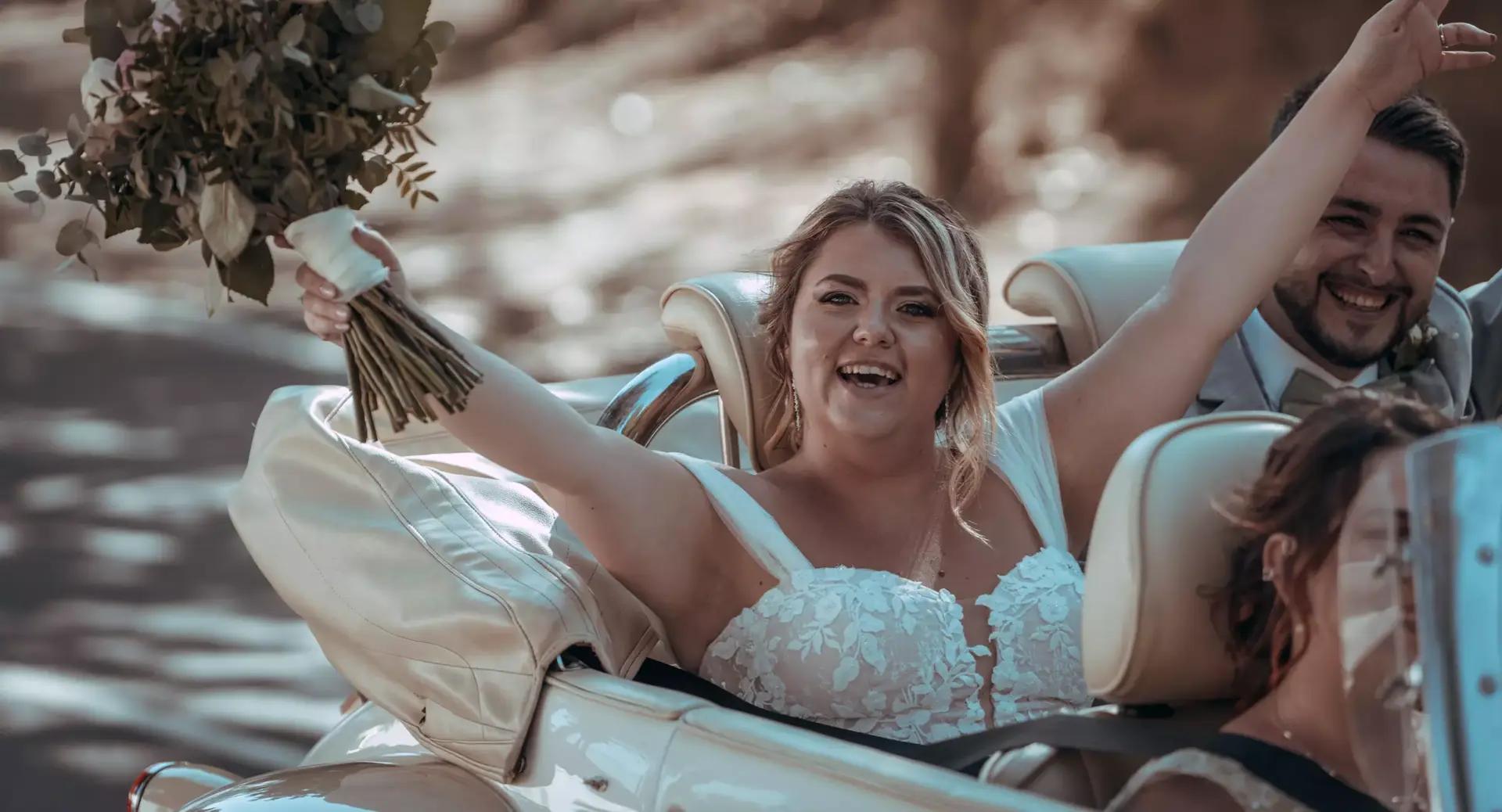 A woman in a wedding dress holds a bouquet and raises her arm, smiling in the front seat of a convertible car, with a man smiling beside her.