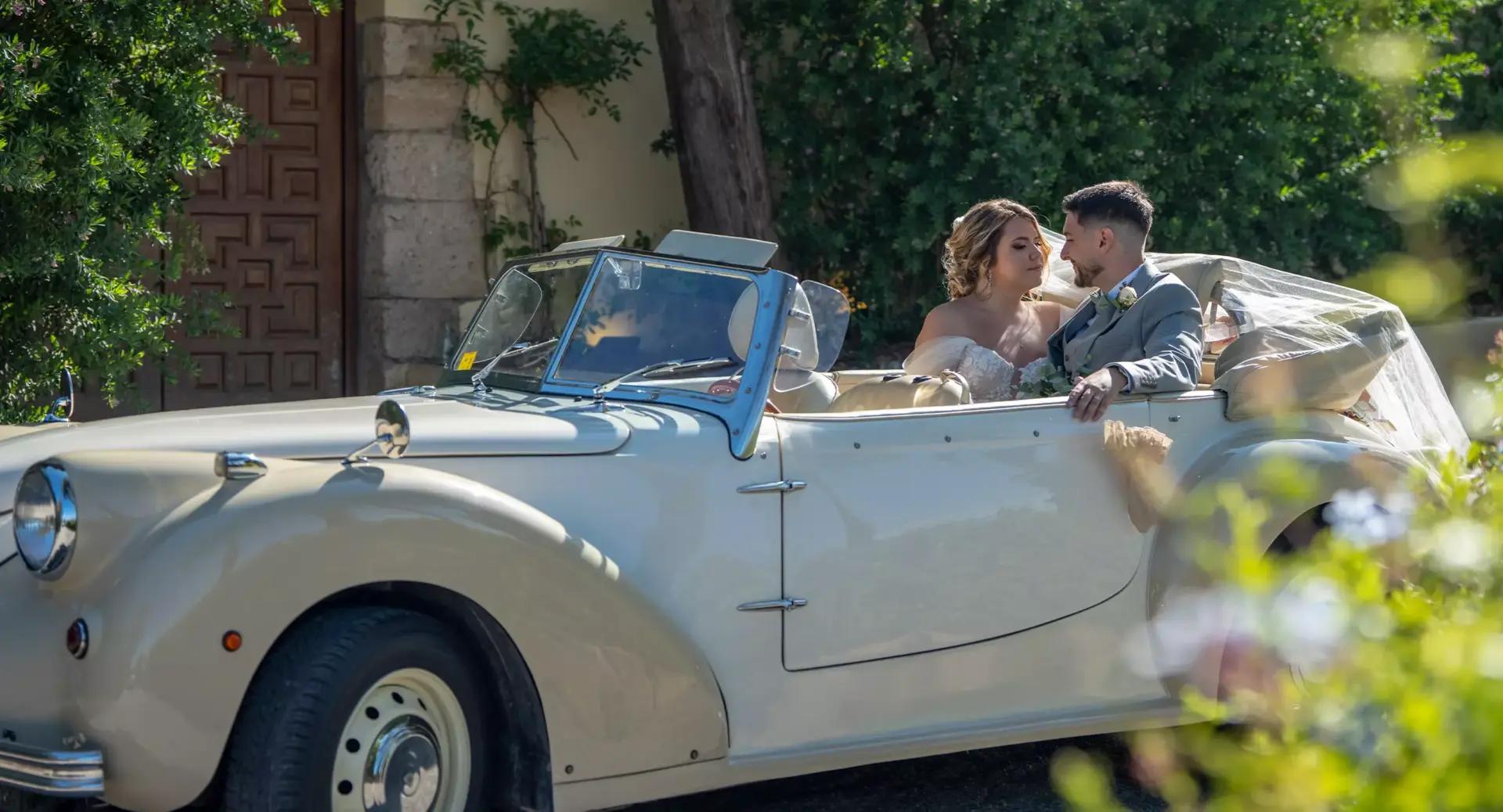 A bride and groom sit together in a vintage white convertible car, dressed in wedding attire, with greenery and a building visible in the background.