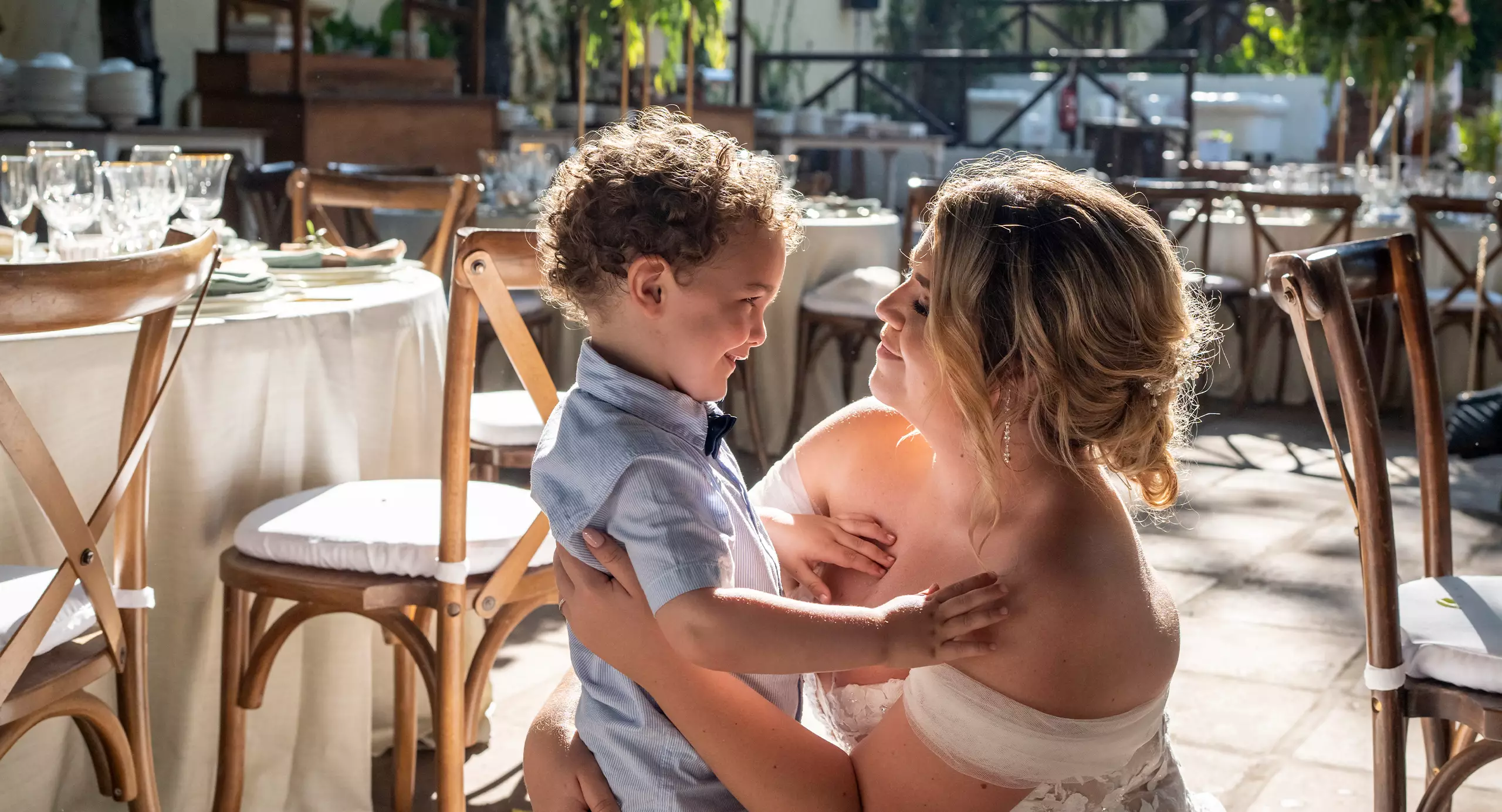 A woman in a white dress kneels and embraces a young boy at an outdoor event with decorated tables and chairs in the background.
