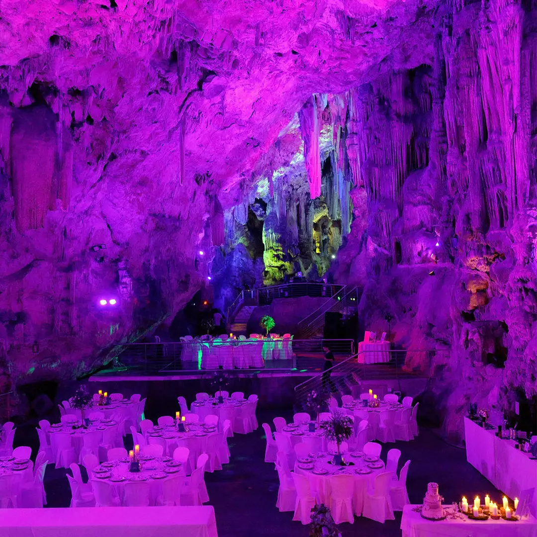 A banquet hall set inside a cave with stalactites, purple lighting, round tables with white tablecloths, and candles arranged for an event.