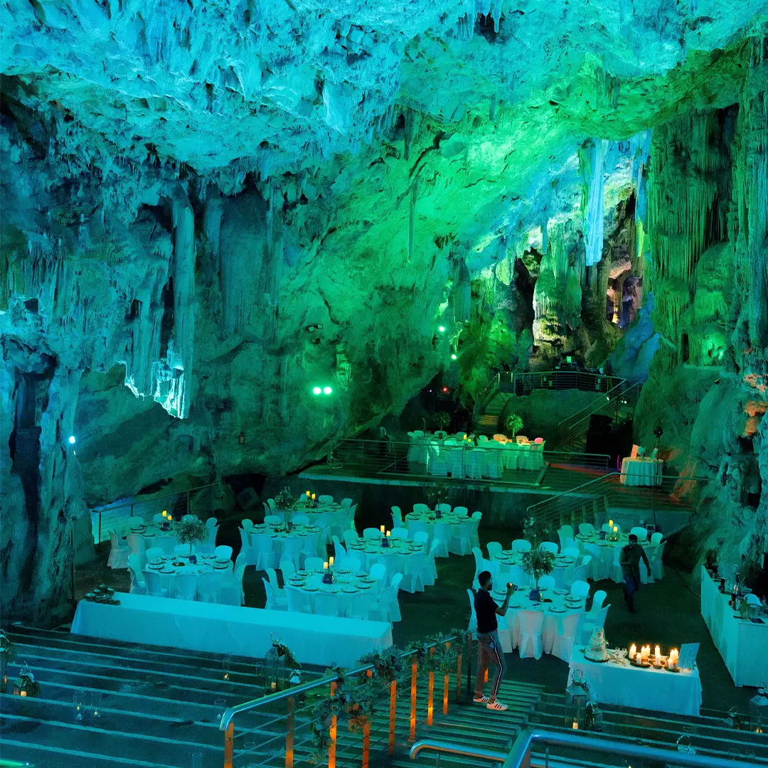 A large underground cave set up as a banquet hall, with white-clothed tables, chairs, and green-blue lighting highlighting the rocky formations.