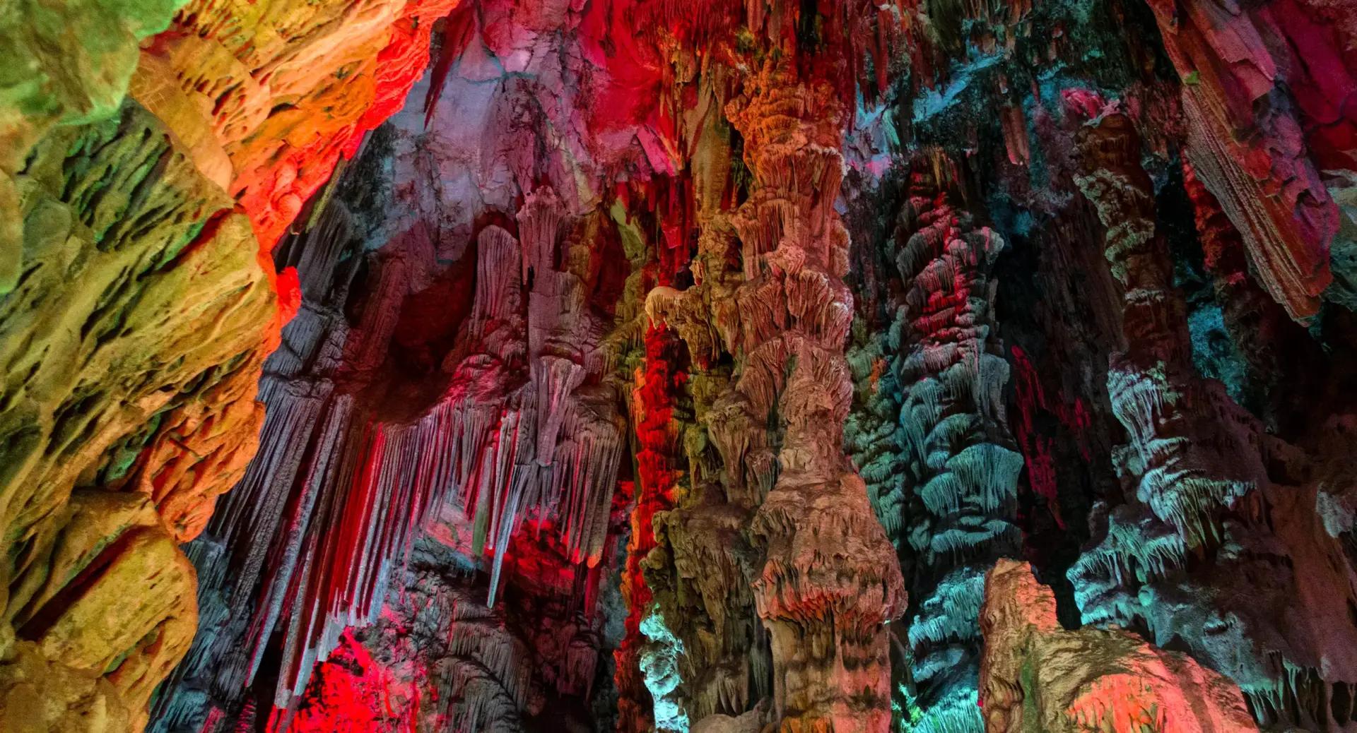 Intricate stalactites and stalagmites inside a cave, illuminated by colorful red, green, and blue lights.