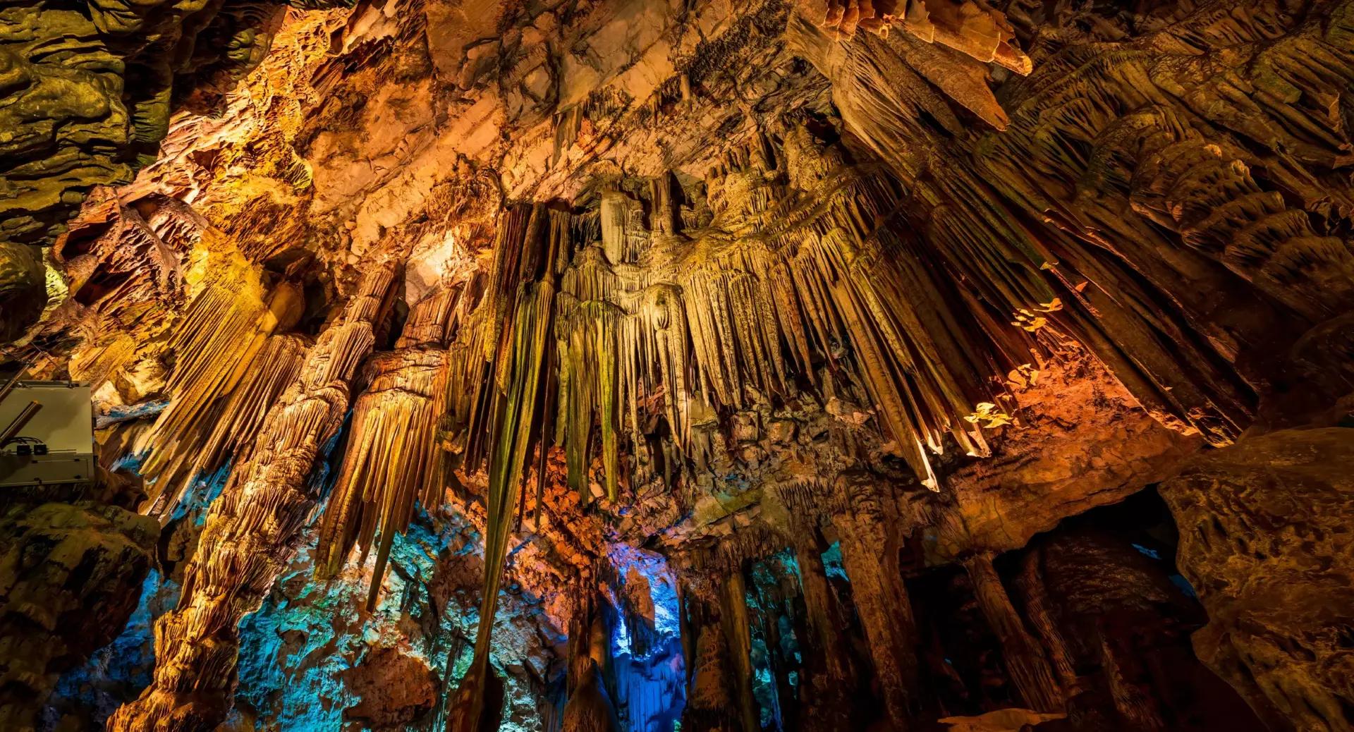 Colorfully lit stalactites and stalagmites hang from the ceiling and rise from the floor inside a large cave chamber.