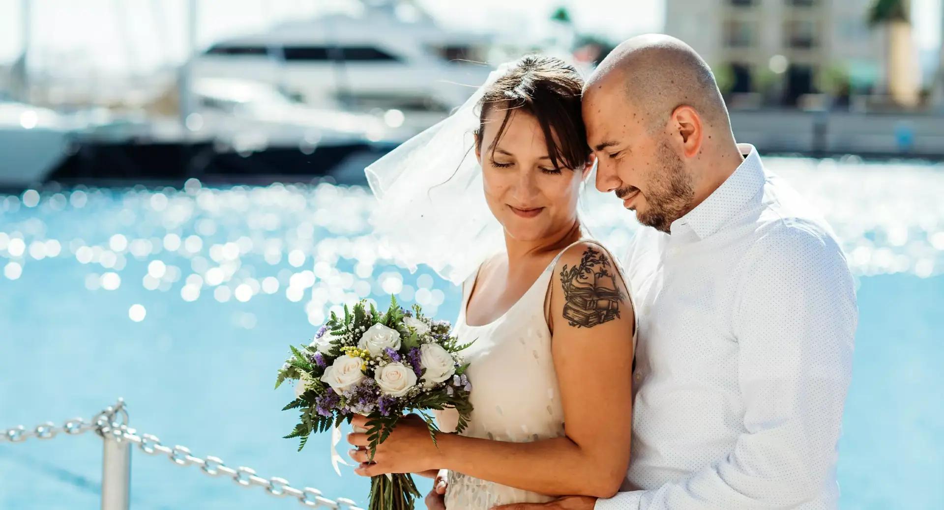 A bride holding a bouquet and a groom stand close together by the water, with boats and sunlight in the background.