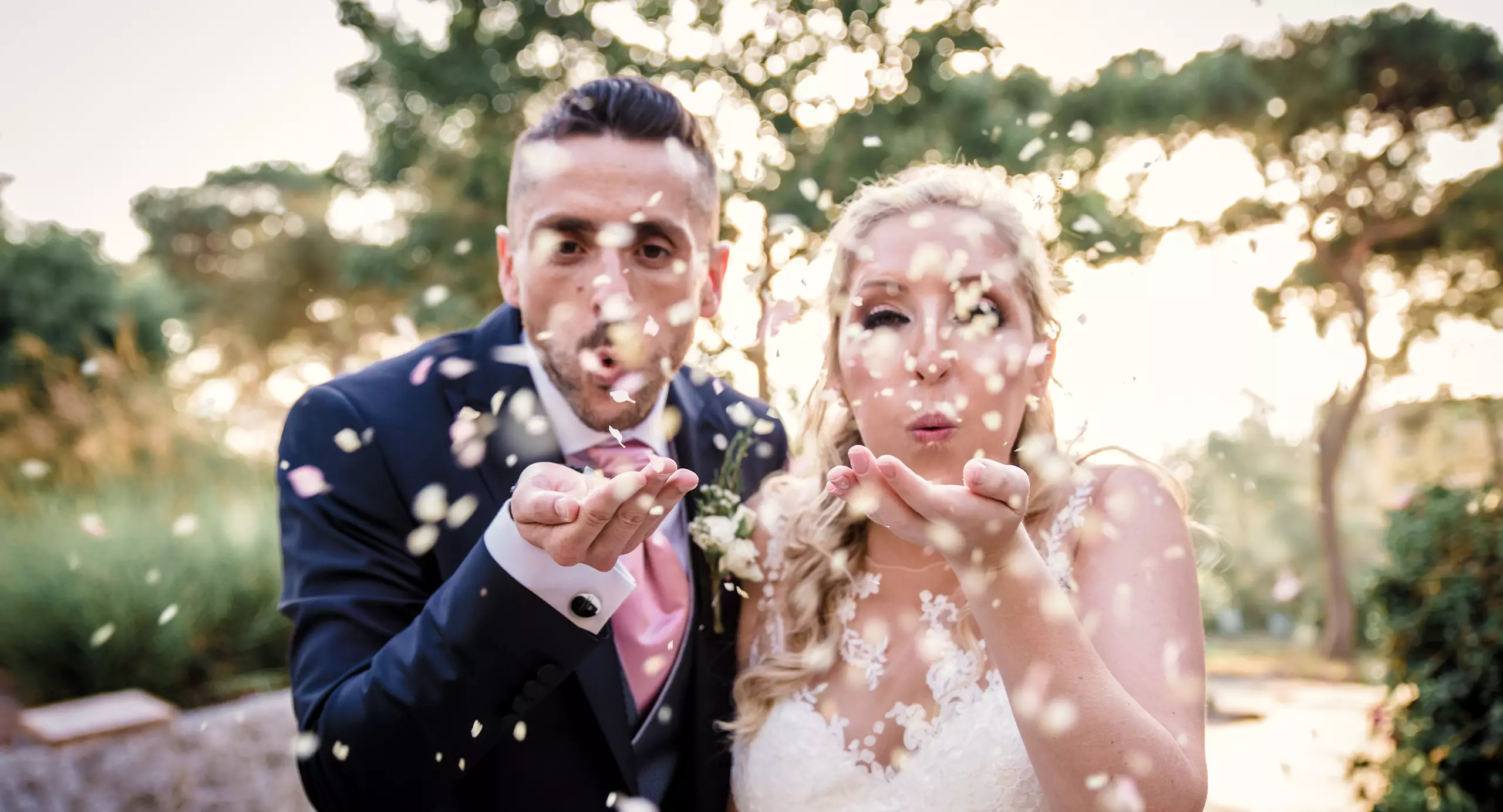 A man in a suit and a woman in a wedding dress blow flower petals toward the camera outdoors, with trees in the background.
