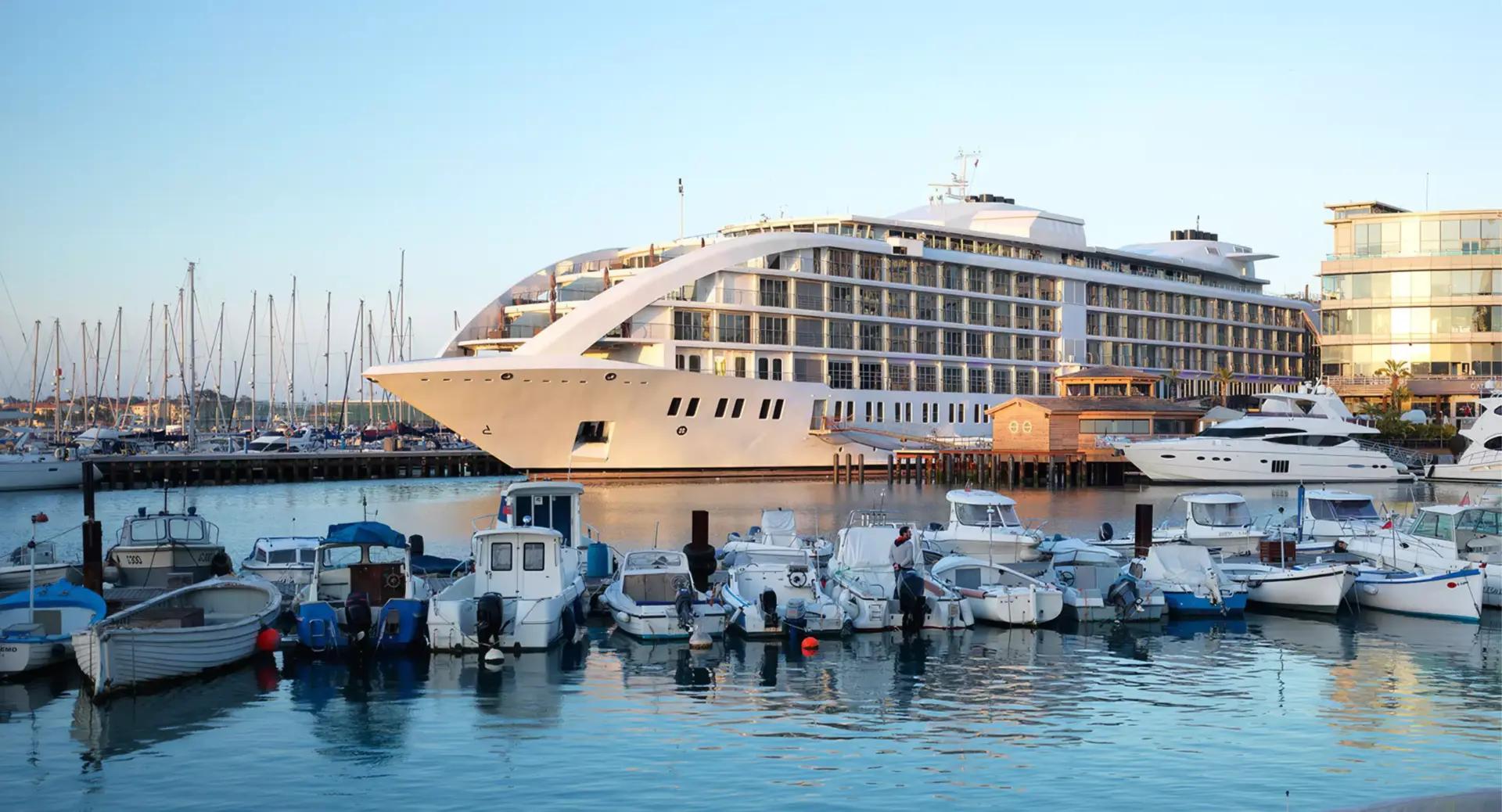A large yacht styled like a cruise ship is docked at a marina with smaller boats moored in the foreground and calm water reflecting the vessels.