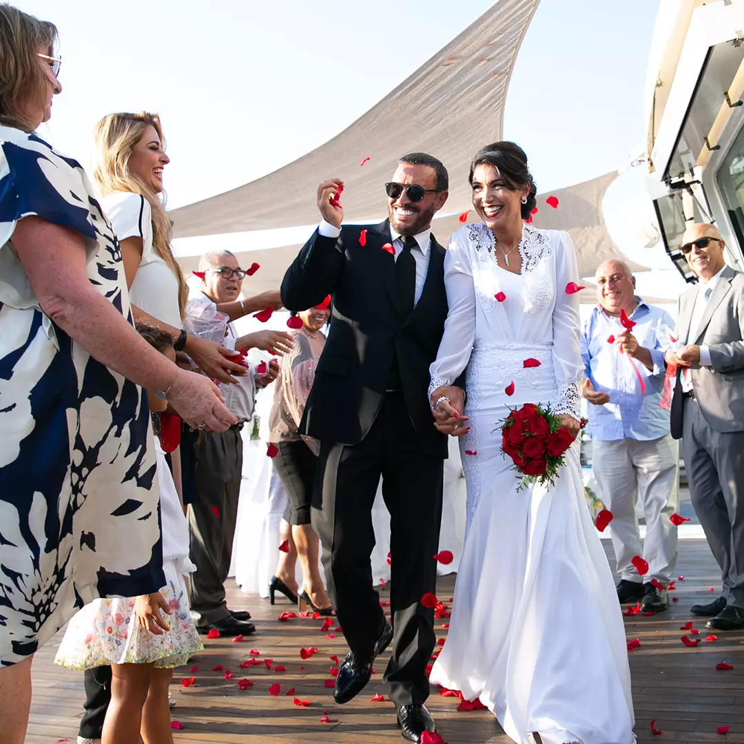 Bride and groom walk down an outdoor aisle, holding hands and smiling, as guests throw red rose petals. The bride holds a bouquet and wears a white dress; the groom wears a black suit.