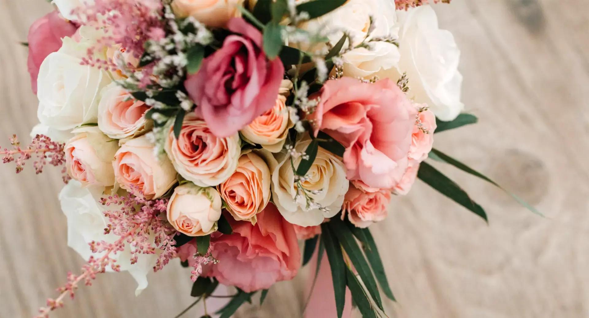 A bouquet of pink, peach, and white roses with greenery, arranged on a light wooden surface.