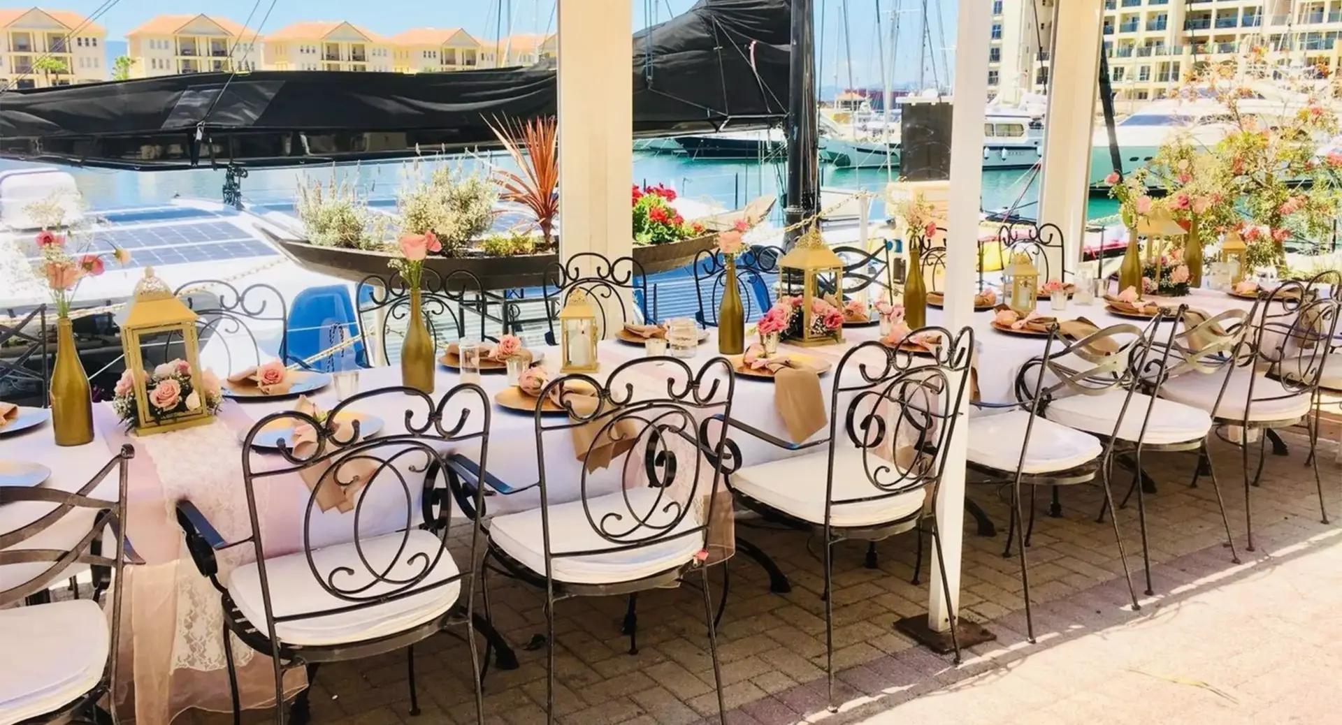 Long outdoor dining table set with floral arrangements, gold accents, and ornate chairs on a patio overlooking boats and water at a marina.