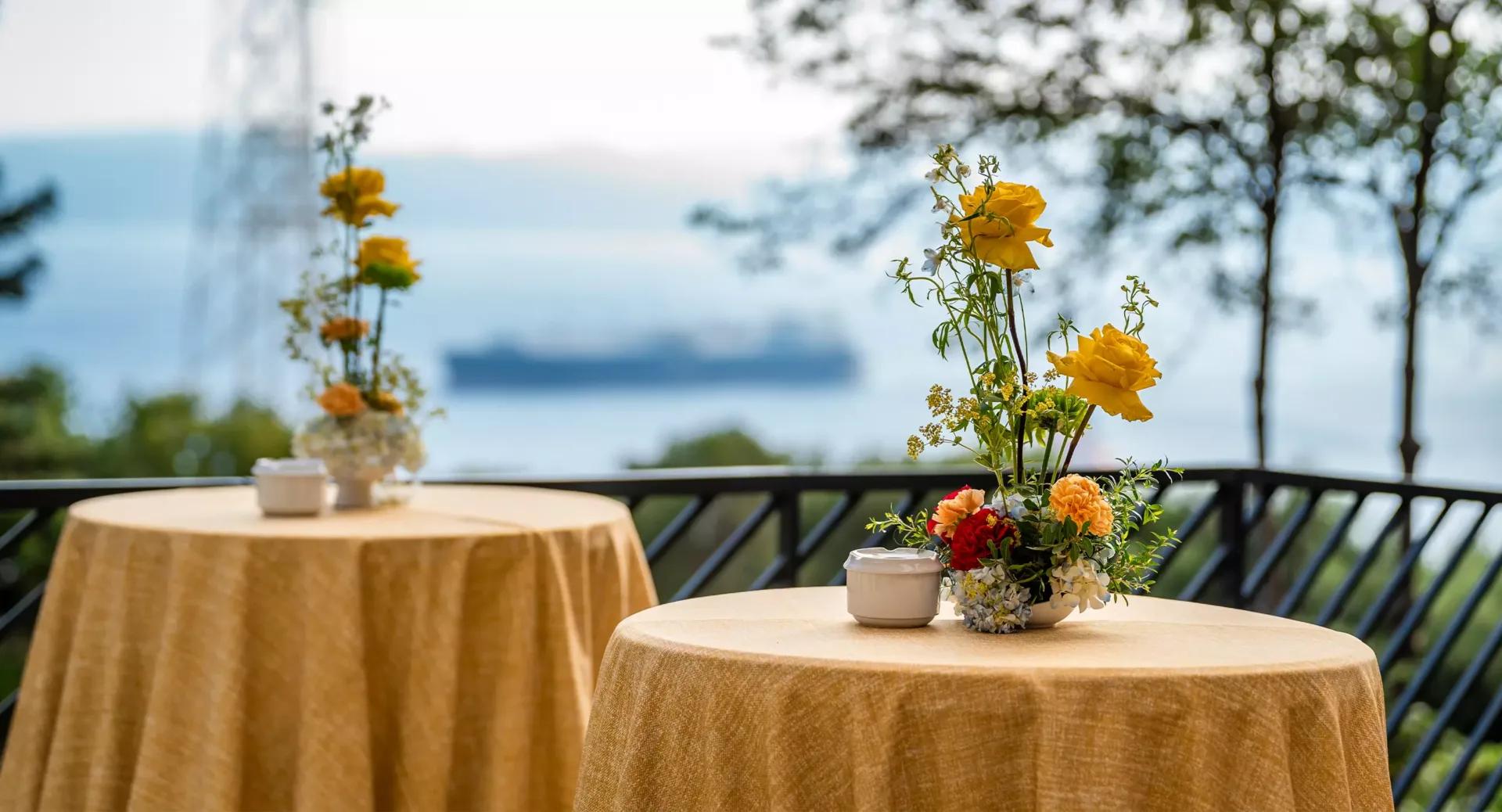 Two round tables with yellow tablecloths and floral centerpieces on a balcony, overlooking water and a distant ship with trees in the background.