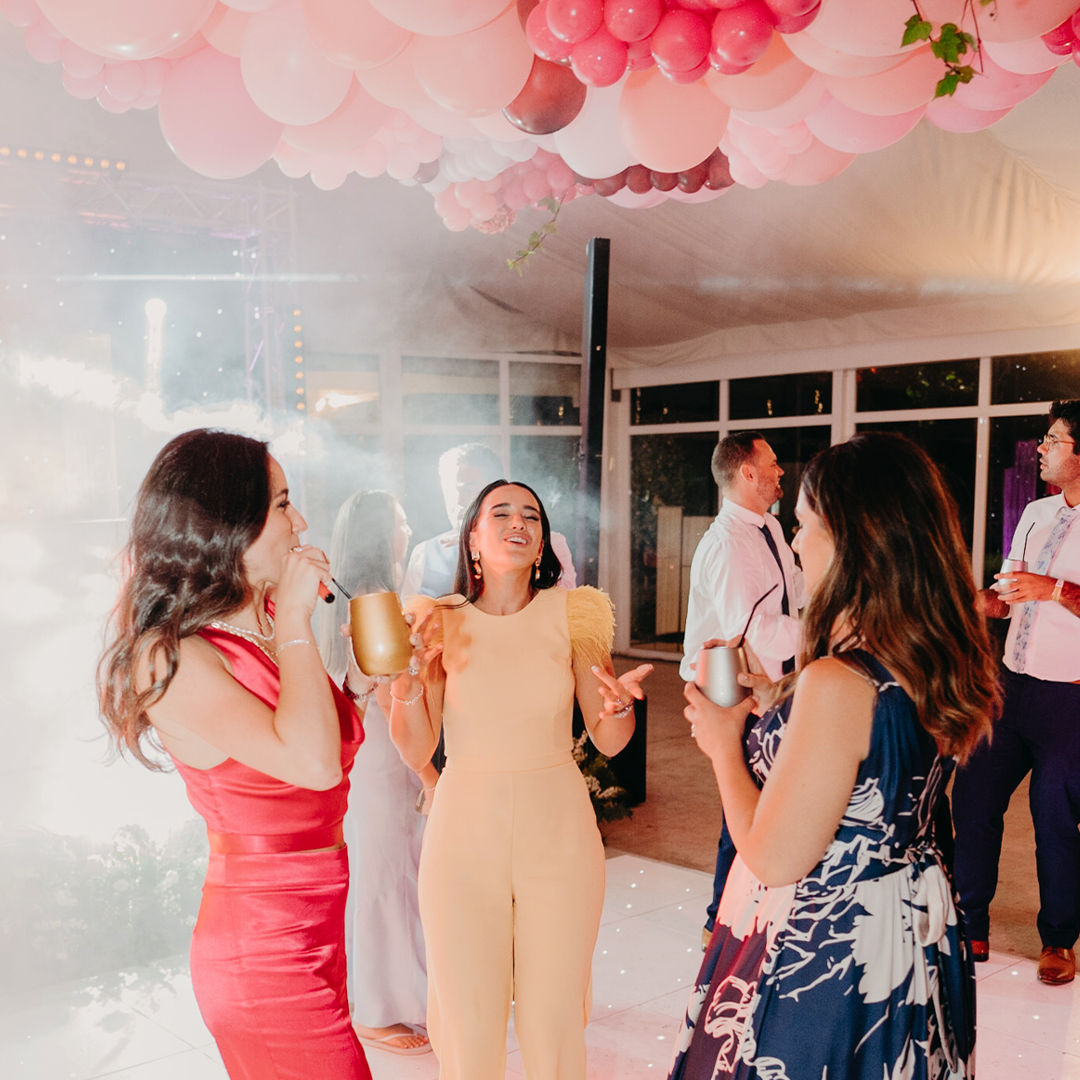 Three women stand and talk on a dance floor under pink balloons at a party, holding drinks, while other guests and smoke effects are visible in the background.