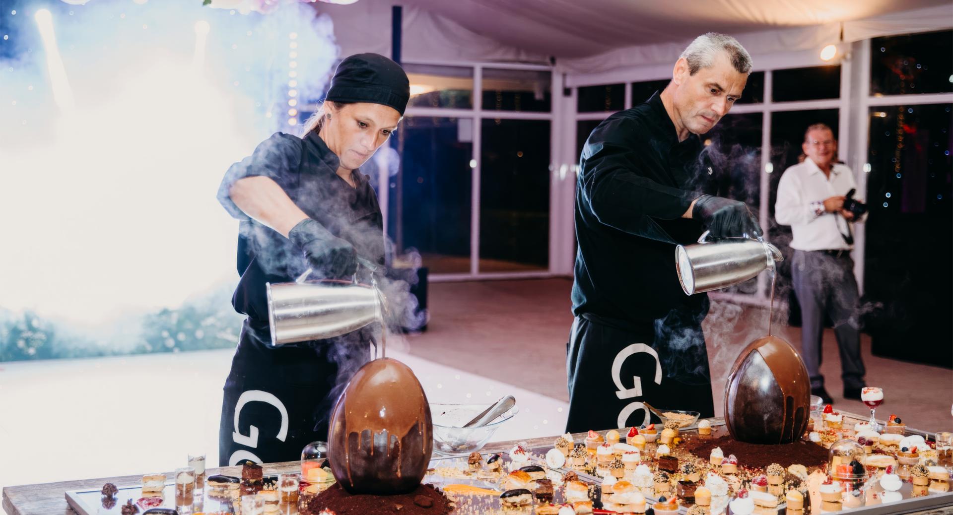Two chefs in black uniforms pour liquid over large chocolate eggs surrounded by desserts on a decorated table, with steam rising and a photographer in the background.
