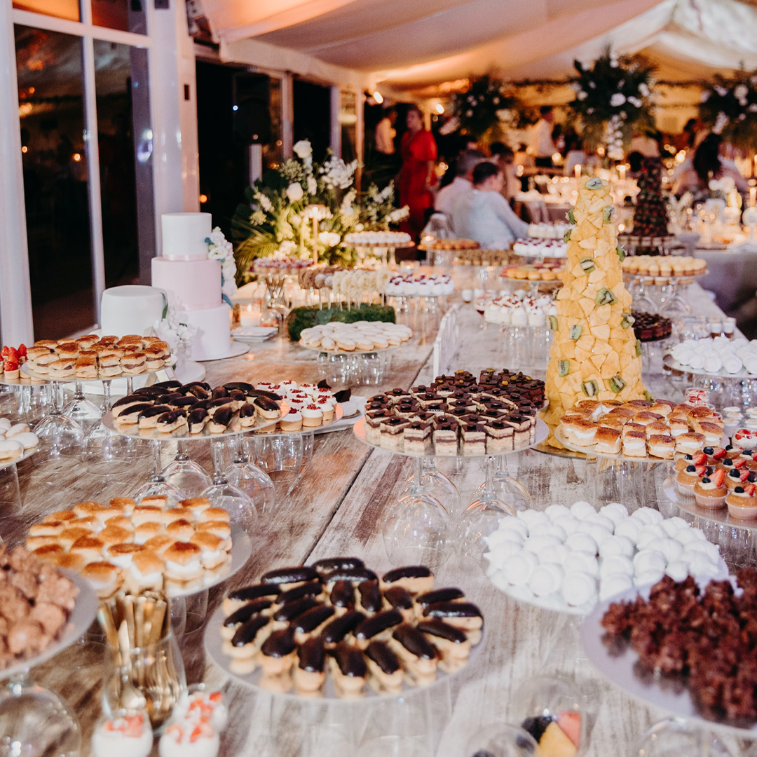 A large dessert table at an event features assorted pastries, cakes, and sweets arranged on glass stands and plates, with floral centerpieces in the background.