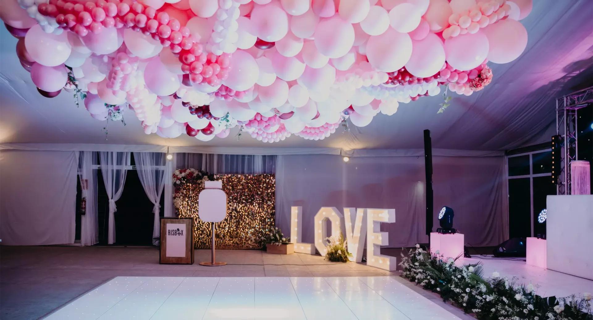 A decorated event space with a ceiling covered in pink and white balloons, illuminated 