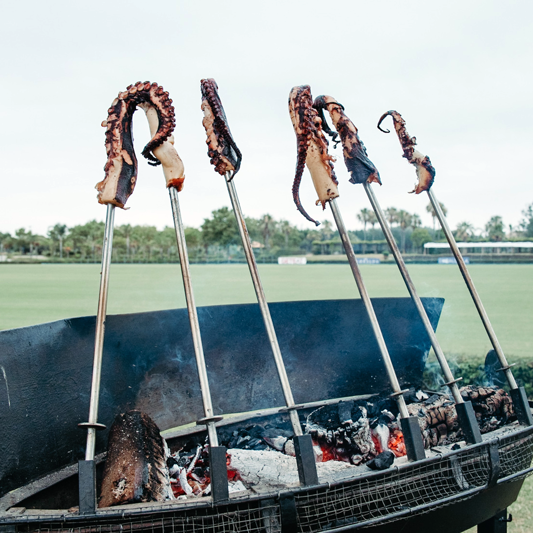 Several skewered octopus tentacles are grilling over an open flame on a large outdoor barbecue with a grassy field in the background.