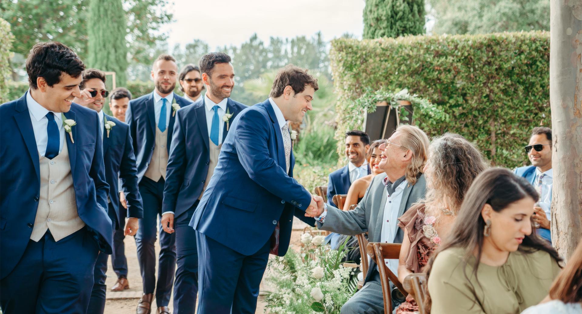 A group of men in suits walk down an outdoor aisle as one man shakes hands with a seated guest among rows of people.