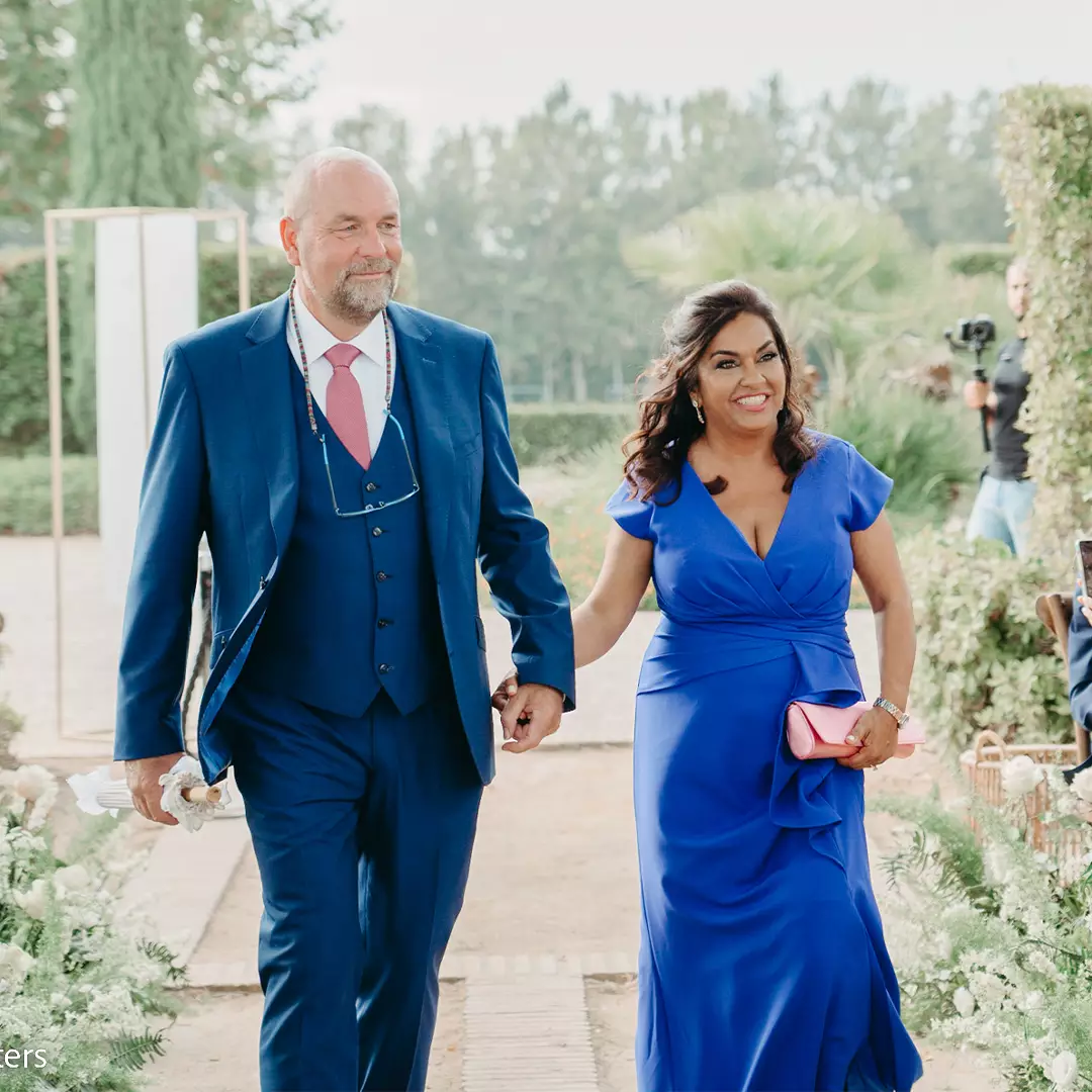 A man and a woman dressed in blue formal attire walk hand in hand outdoors at what appears to be a wedding or formal event.