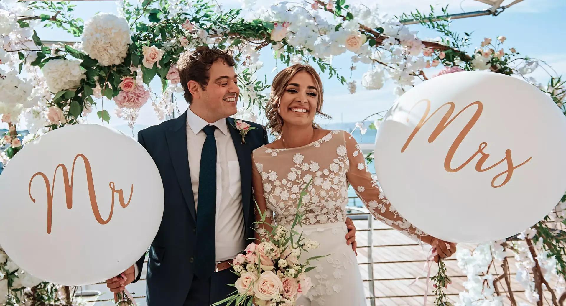 A bride and groom stand smiling under a floral arch, holding large white balloons labeled 