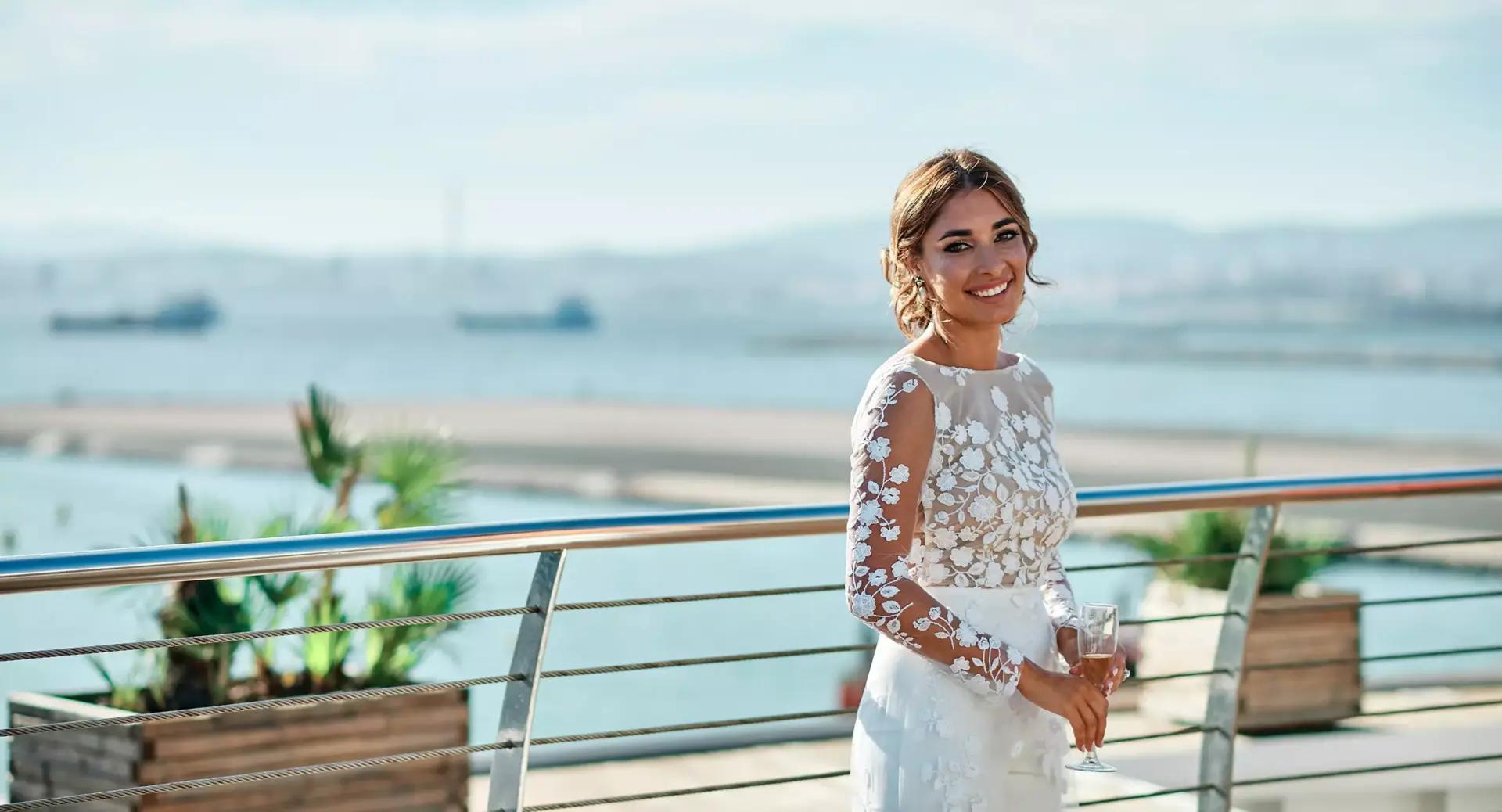 A woman in a white lace dress stands by a railing, holding a glass and smiling, with a waterfront and ships visible in the background.
