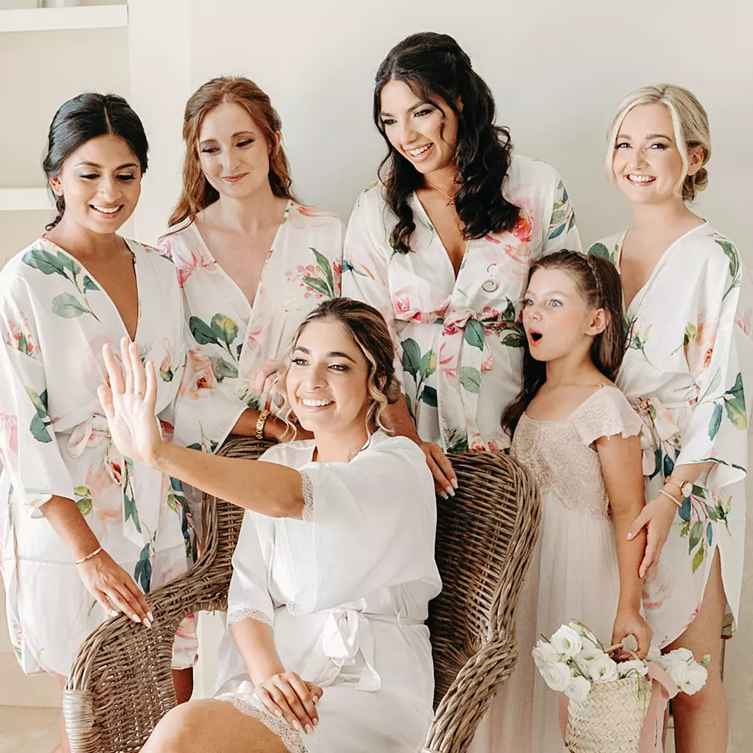 A bride seated in a wicker chair shows her ring to her bridesmaids and a young flower girl, all wearing floral robes, indoors.