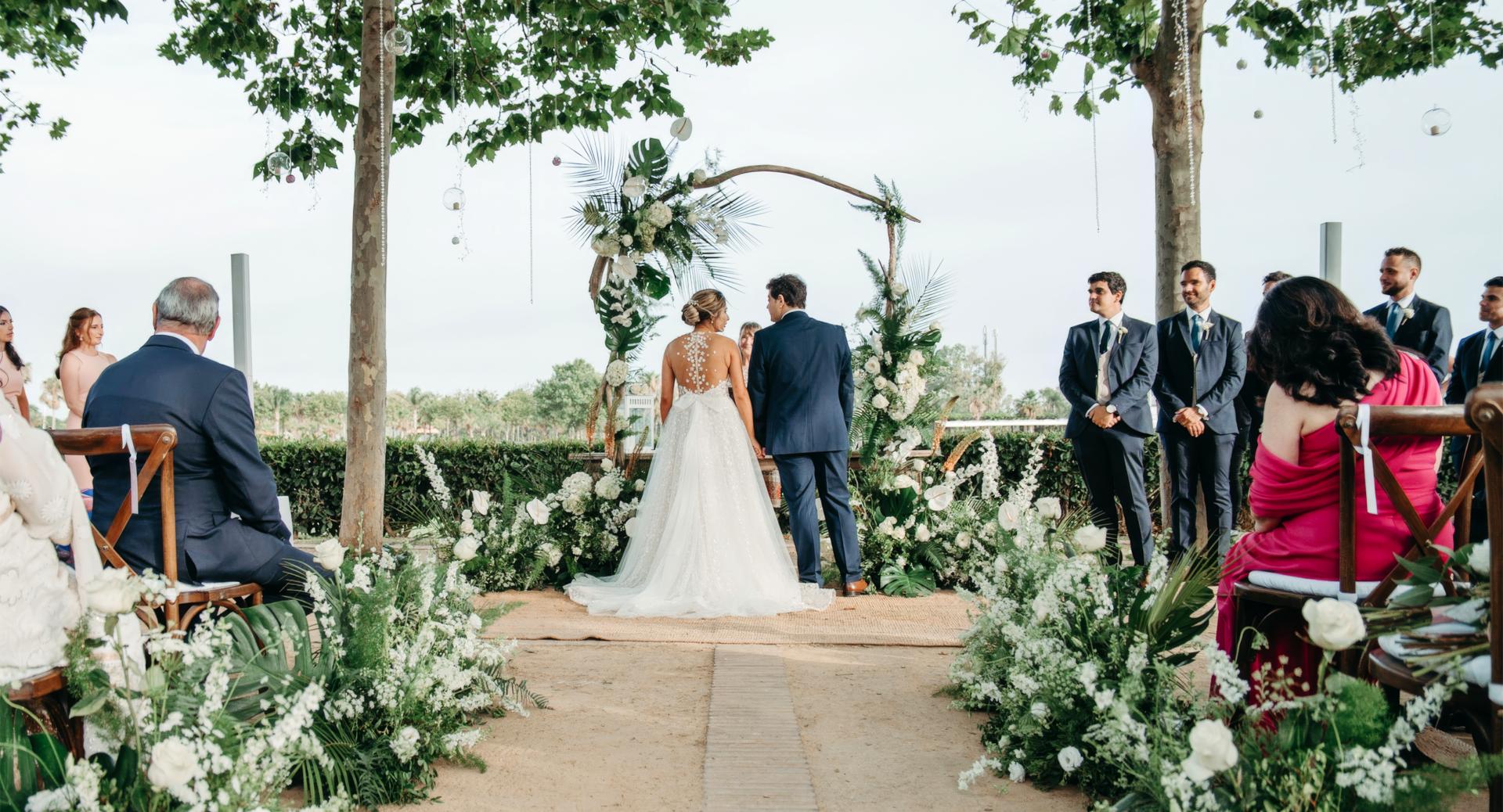 A bride and groom stand together at an outdoor wedding ceremony, surrounded by guests seated on both sides and decorated with white flowers and greenery.