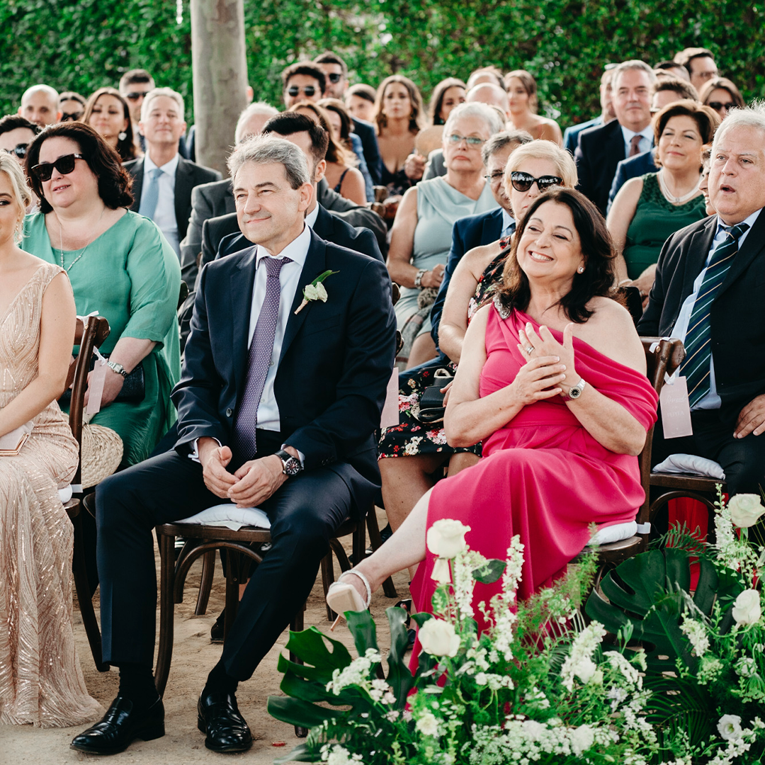 Guests seated outdoors at a wedding ceremony, dressed in formal attire, with greenery and floral decorations visible in the foreground.