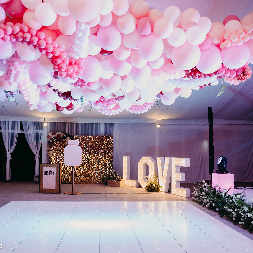 A decorated event space with a white floor, pink and white balloon ceiling, a gold sequin backdrop, and large illuminated letters spelling 