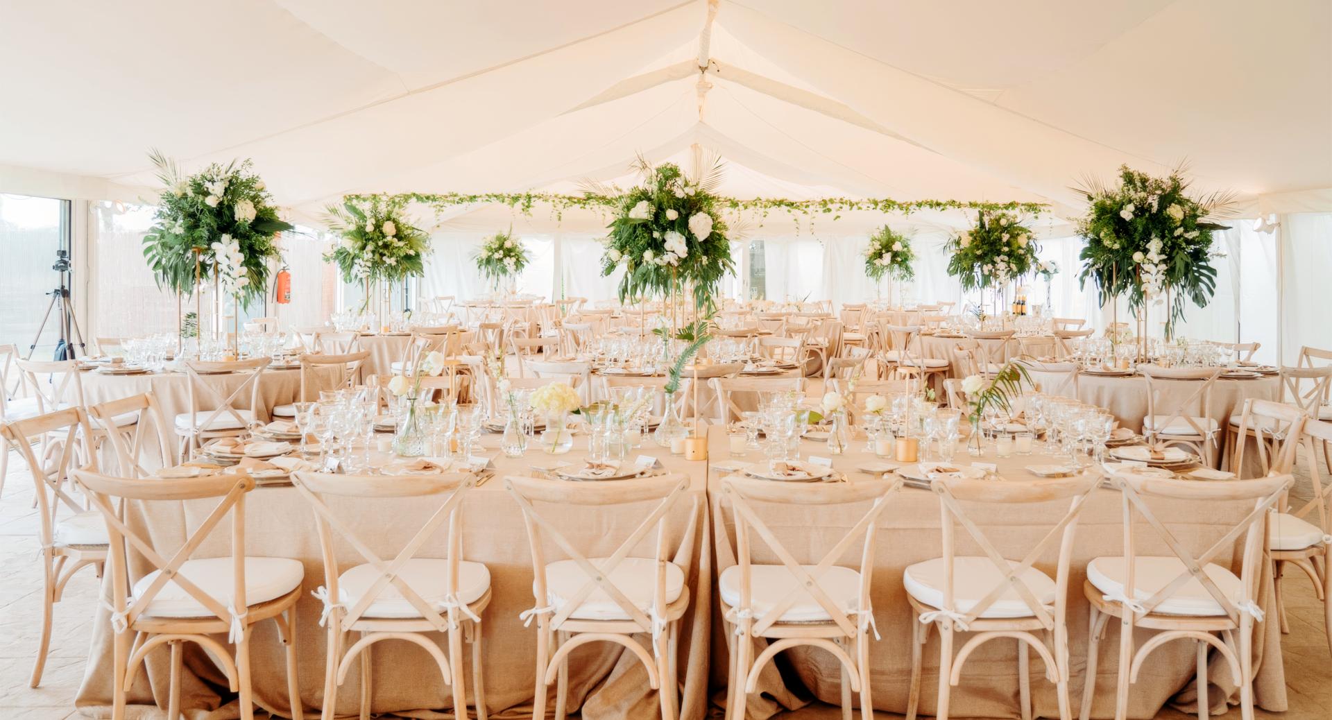 Elegant event tent with long banquet tables set for dining, decorated with tall floral centerpieces and neatly arranged wooden chairs.