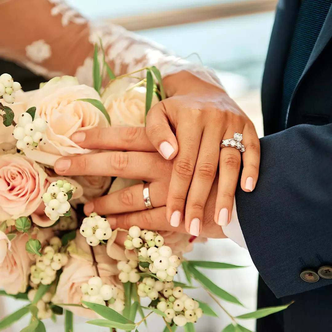 Close-up of a bride and groom’s hands with wedding rings, resting on a bouquet of pale roses and white berries.