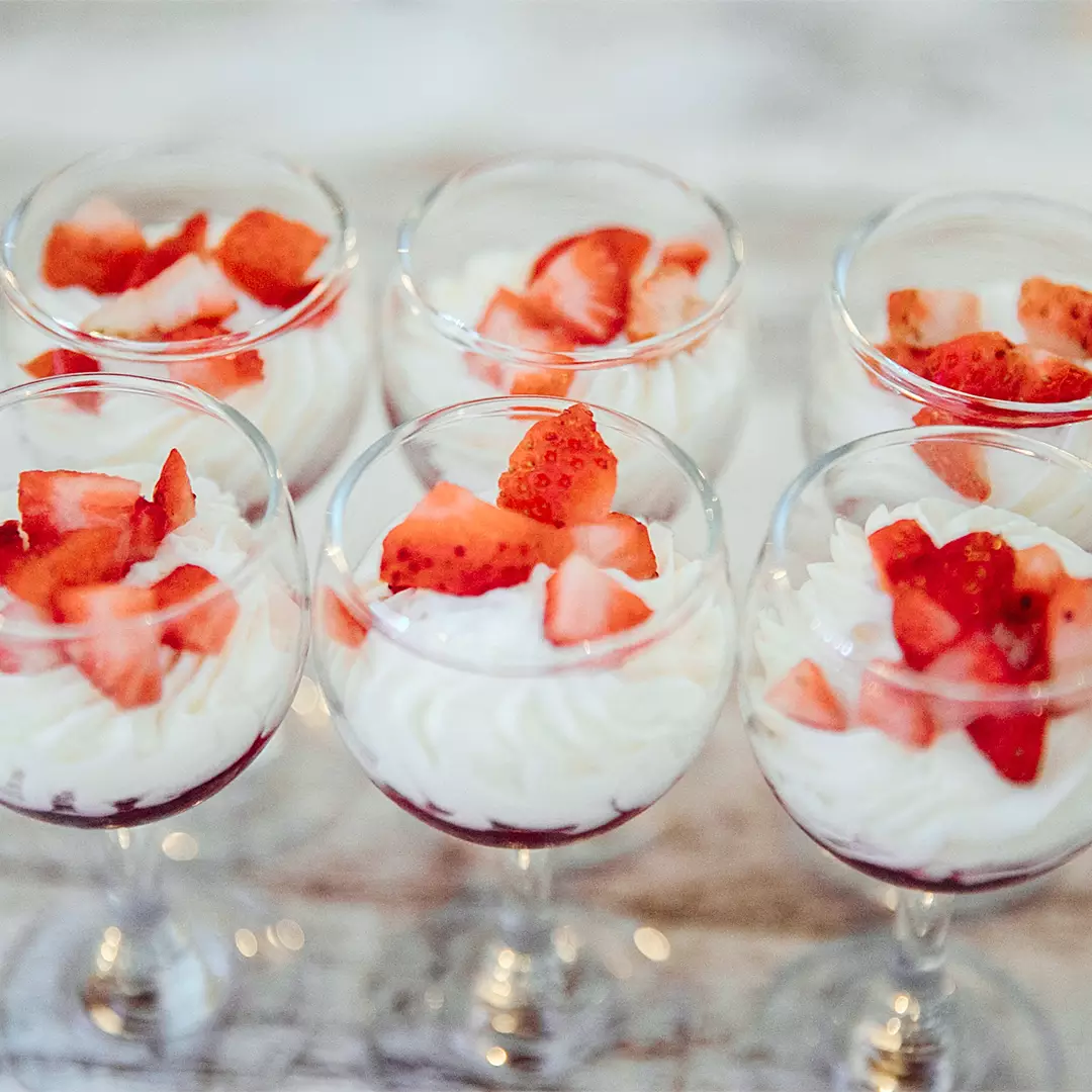 Six glass cups filled with swirled whipped cream topped with chopped fresh strawberries, arranged together on a light-colored surface.