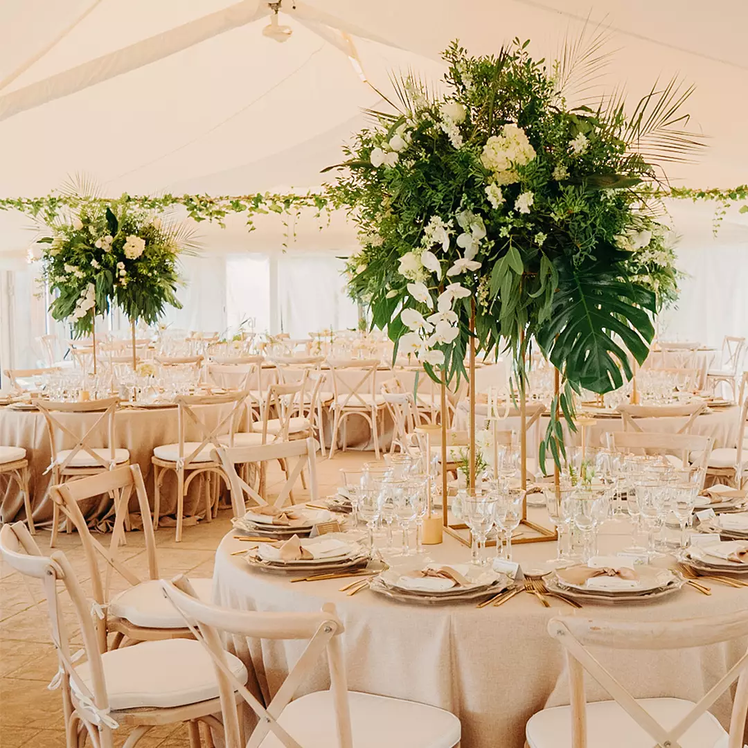 Elegant banquet hall with round tables set for an event, featuring tall floral centerpieces with greenery and white flowers under a light-filled tent.