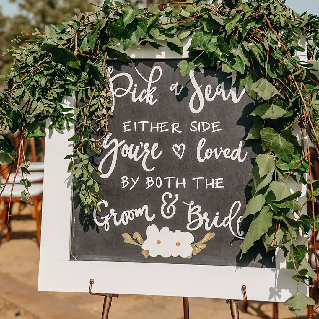 A framed chalkboard wedding sign with greenery reads, 