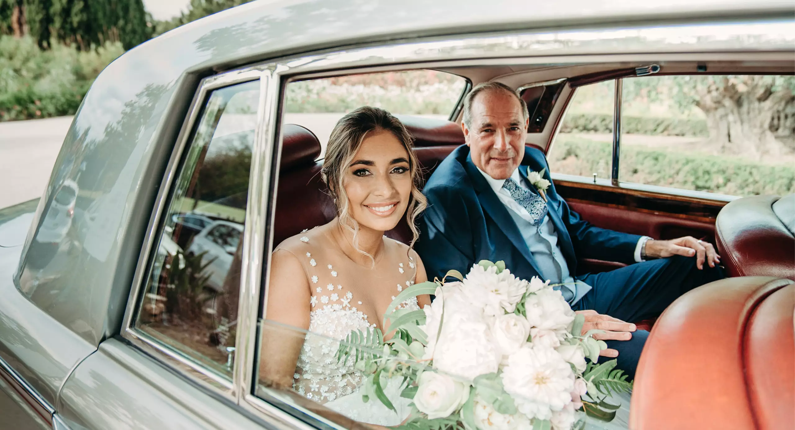 A bride in a white dress holding a bouquet sits in the back of a car smiling, with a man in a suit beside her.