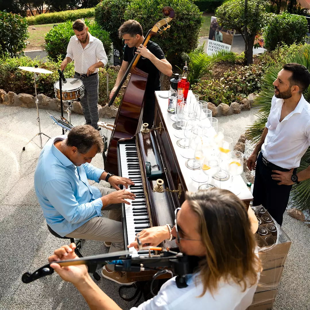 Five men play instruments and gather around an outdoor piano bar setup with drinks; the group includes a pianist, drummer, bassist, violinist, and one man standing nearby.