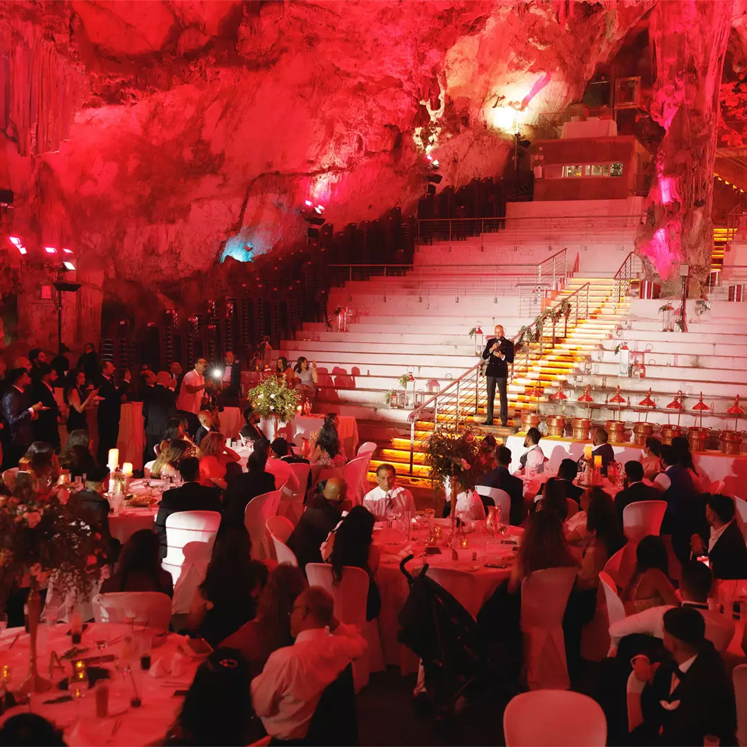 A formal event with seated guests is held in a large cave, featuring red lighting, white tablecloths, and a speaker standing on illuminated stairs.