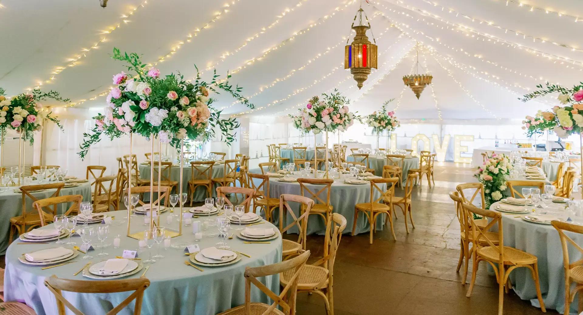 Elegant wedding reception setup inside a tent with round tables, floral centerpieces, wooden chairs, string lights, and a large illuminated 