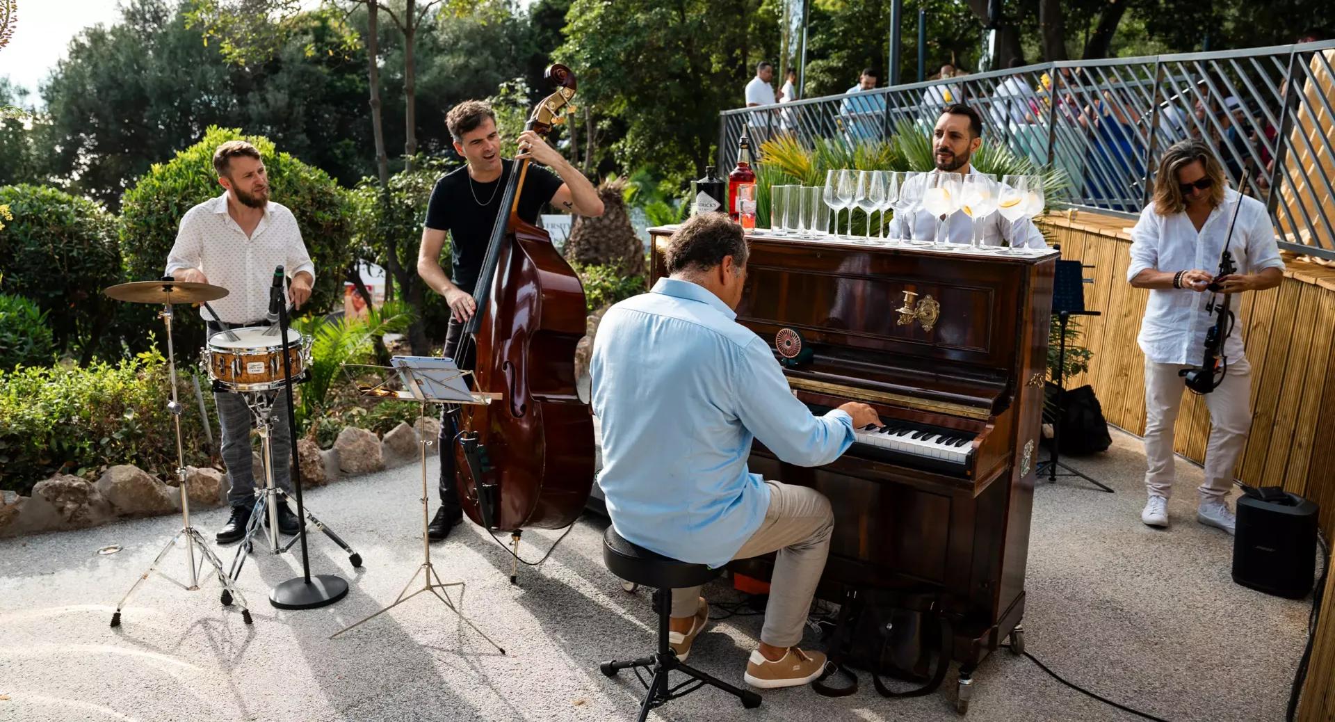 A jazz band plays outdoors. A pianist sits at a piano while a bassist, drummer, and violinist stand nearby.